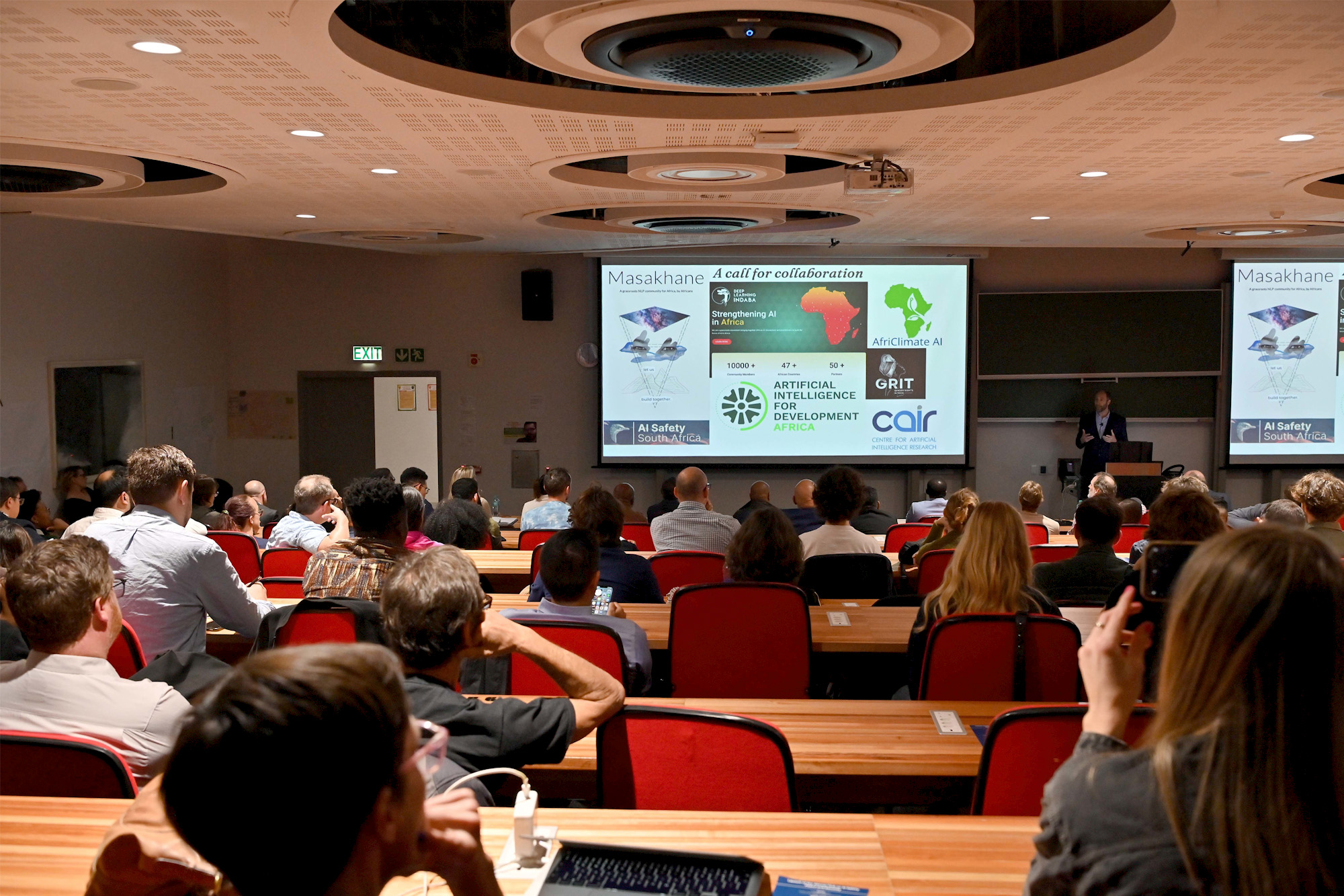 Delegates attend the University of Cape Towns launch | Brimco Audience attending the university of cape town event launching the african hub on ai safety peace security with a presenter speaking before a large projected presentation on ai collaboration in africa