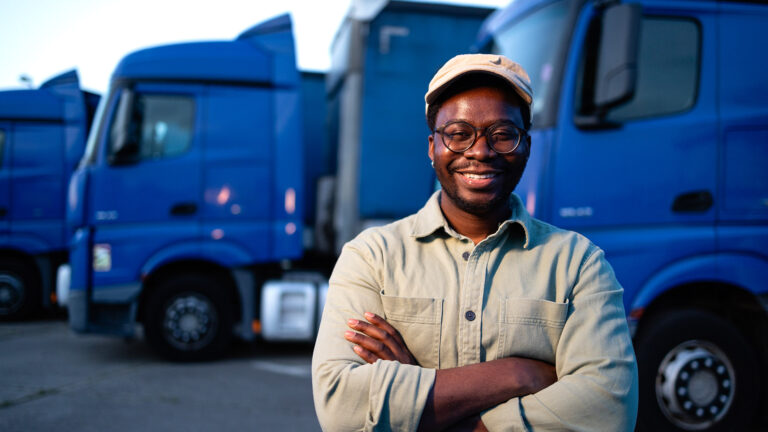 Smiling african logistics manager or truck driver standing confidently in front of a row of blue commercial trucks