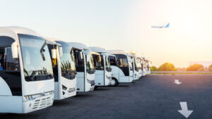 Row of white airport shuttle buses parked at sunrise with an airplane flying in the background