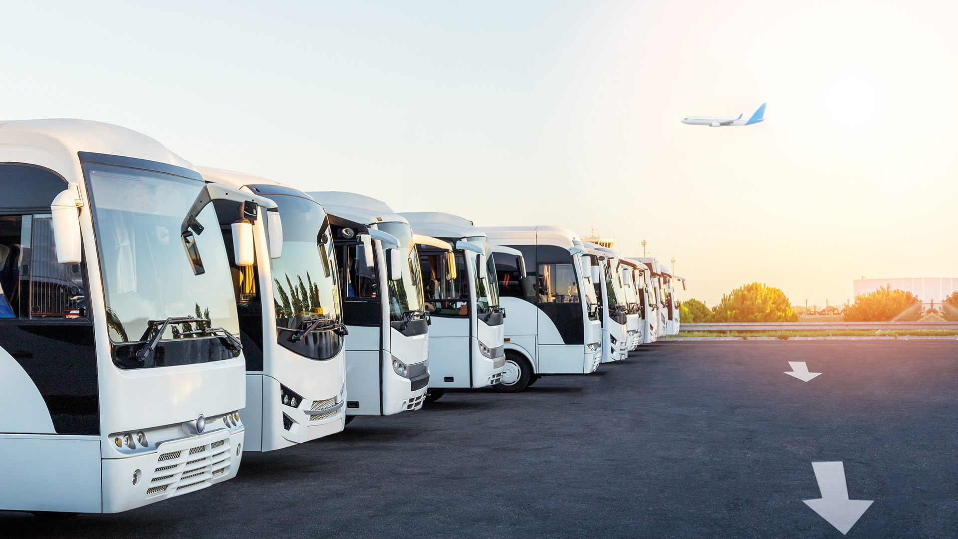 Row of white airport shuttle buses parked at sunrise with an airplane flying in the background