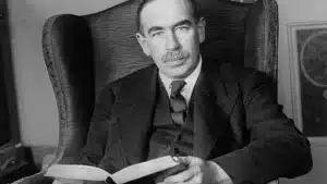 Black and white photo of john maynard keynes sitting in an armchair holding an open book and looking directly at the camera