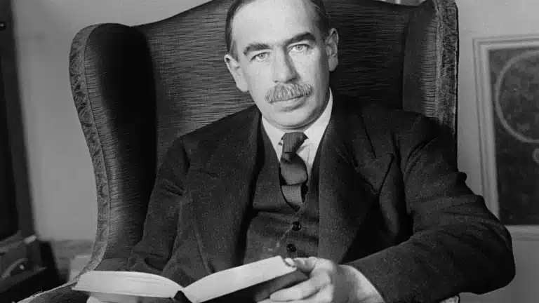 Black and white photo of John Maynard Keynes sitting in an armchair, holding an open book and looking directly at the camera.