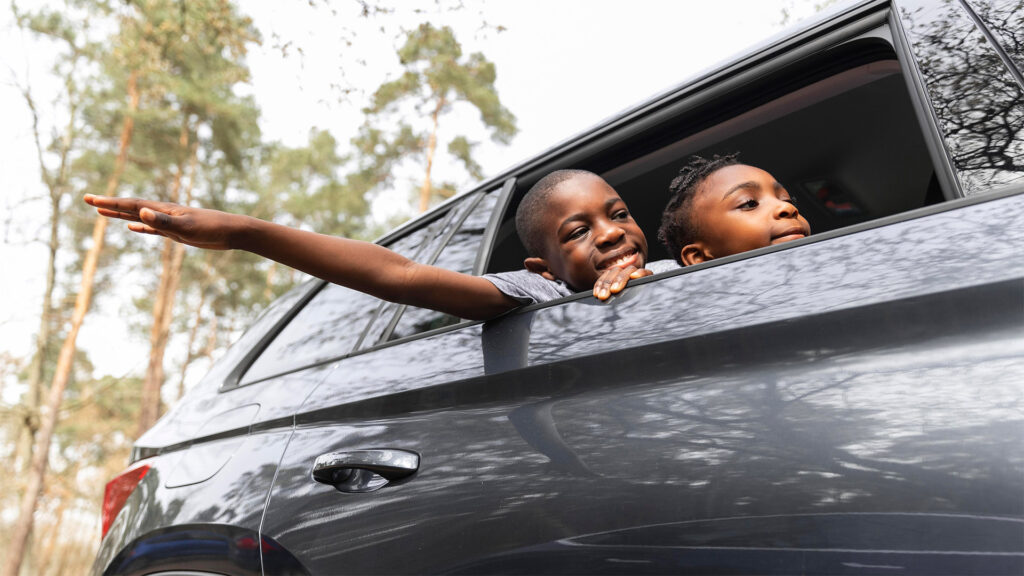 Two happy African children leaning out of a car window, smiling and enjoying the ride through a forested area.