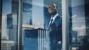 African businessman in a suit looking through an office window with city skyscrapers in the background symbolizing ambition and strategic vision