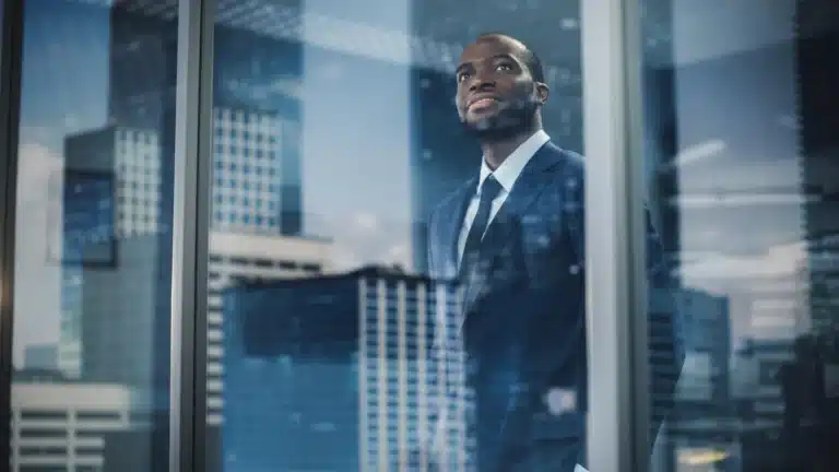 African businessman in a suit looking through an office window with city skyscrapers in the background, symbolizing ambition and strategic vision.