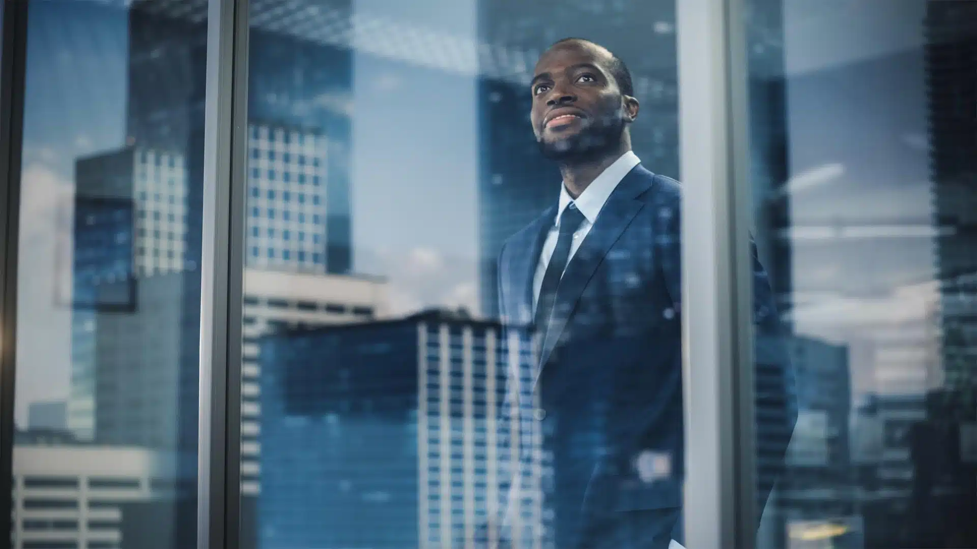 African businessman in a suit looking through an office window with city skyscrapers in the background symbolizing ambition and strategic vision