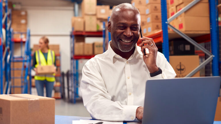 Smiling warehouse manager talking on the phone while working on a laptop at a packing station surrounded by cardboard boxes