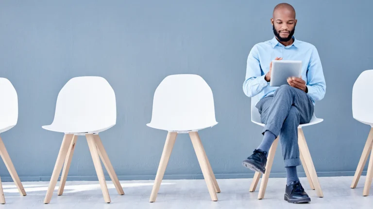 A man sitting in a waiting area using a tablet, symbolizing job applications and employment background screening.