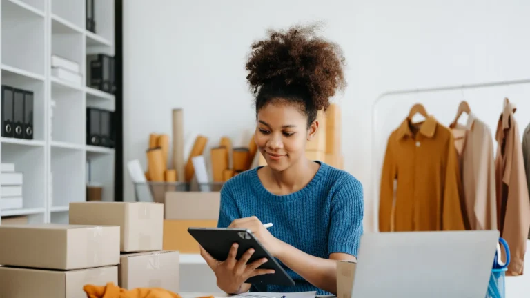 A small business owner prepares online orders, using a tablet while surrounded by shipping boxes.