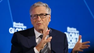 Bill gates speaking at a global business forum against a blue background gesturing with his hands while addressing the audience