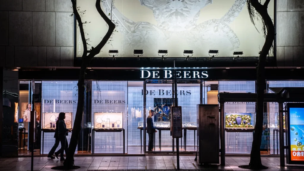 Nighttime exterior of a de beers jewelry store with lit window displays and pedestrians walking past