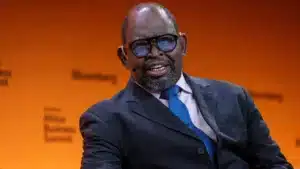 A man wearing a dark suit blue tie and glasses speaks on stage against a yellow backdrop displaying the bloomberg africa business summit branding