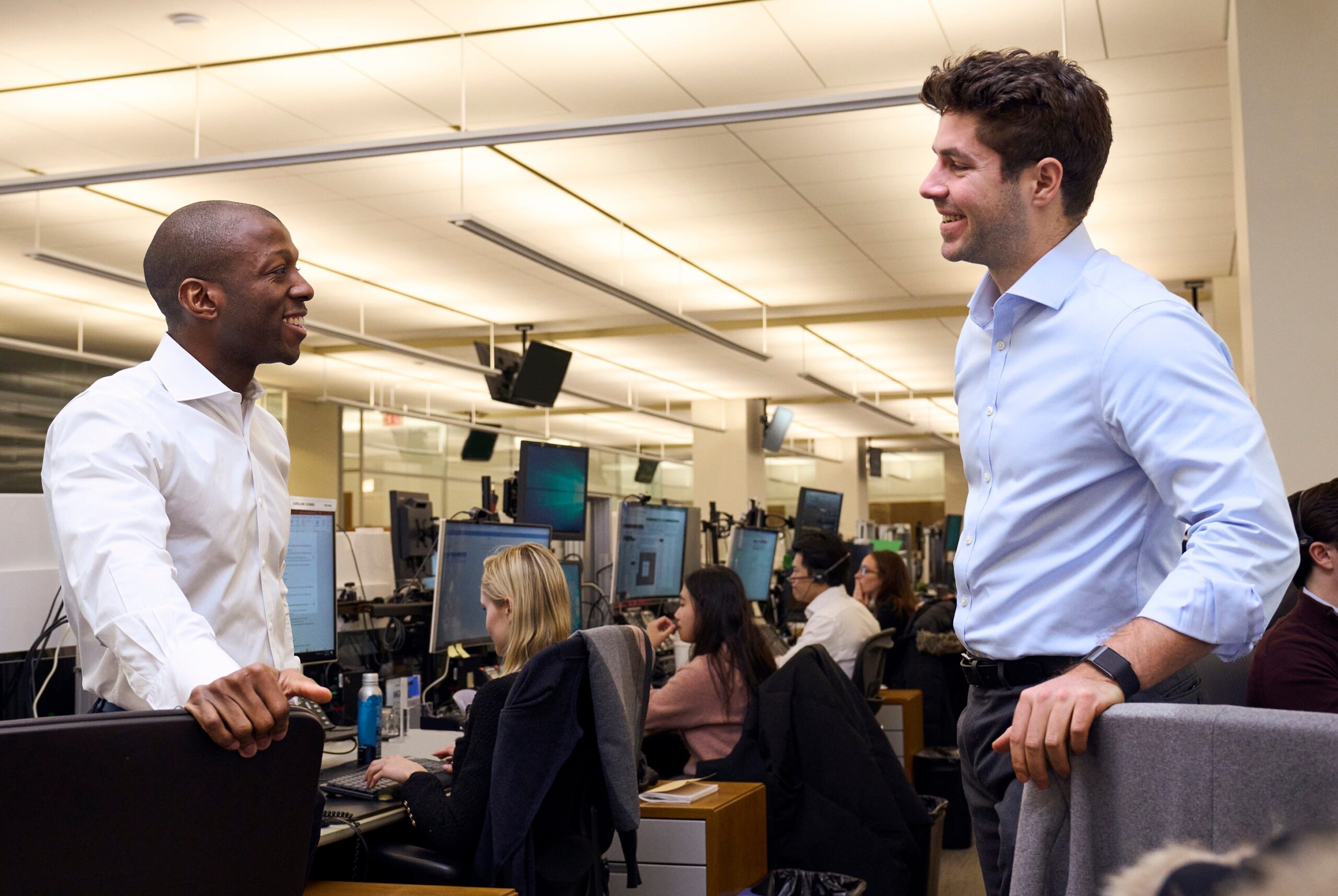 Two goldman sachs employees smiling and conversing on the trading floor with other colleagues working at computer stations in the background