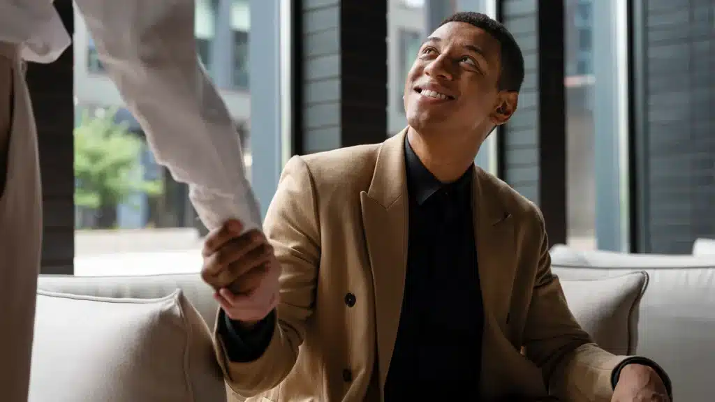 A seated man in a tan suit smiles warmly as he shakes hands with another person in a light-colored shirt inside a modern office setting.