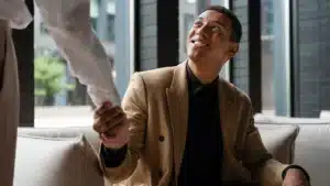 A seated man in a tan suit smiles warmly as he shakes hands with another person in a light colored shirt inside a modern office setting