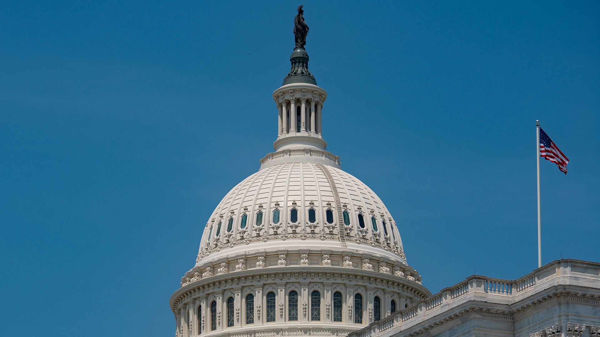 The United States Capitol Dome Under Clear Skies | Brimco The dome of the u S Capitol building with an american flag waving nearby against a clear blue sky