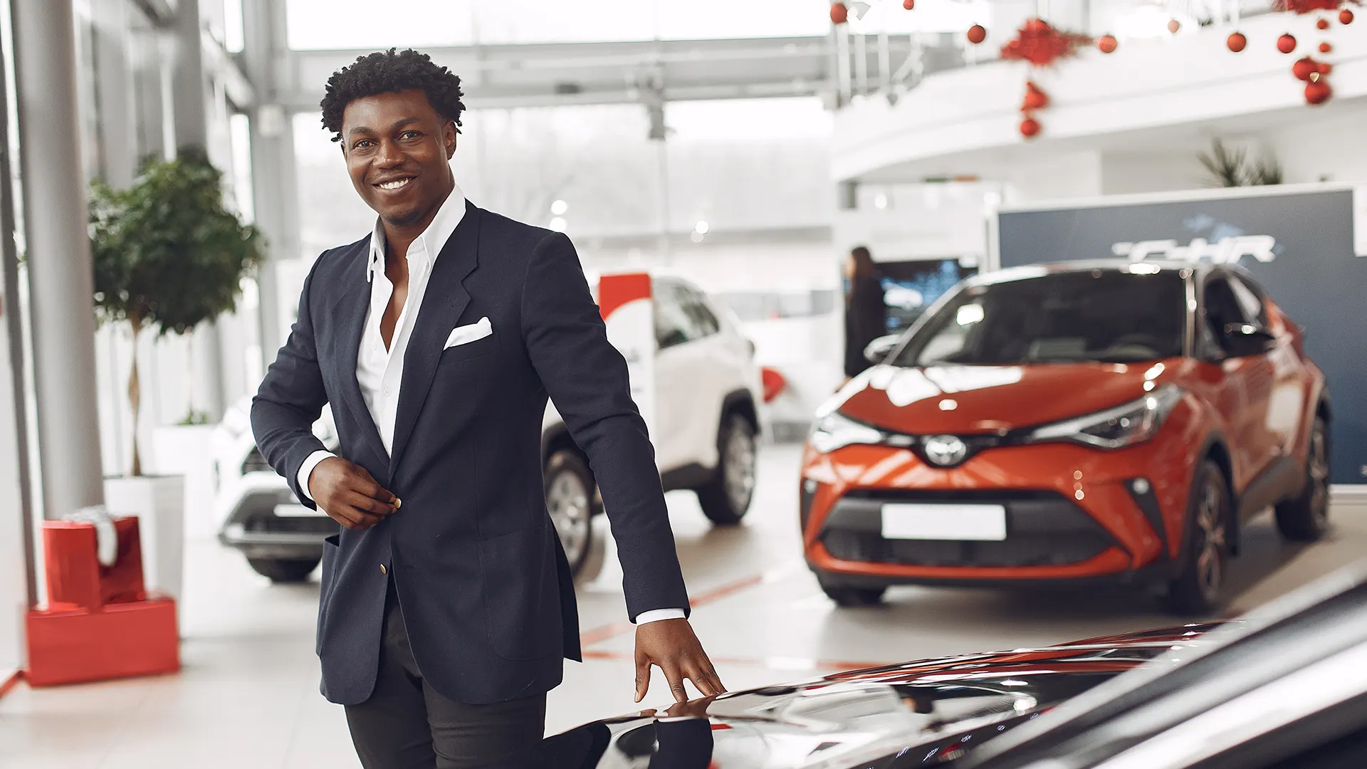 Car Sales Agent in Showroom | Brimco Smiling car sales agent standing in a dealership showroom with new vehicles behind him