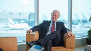 Sir tim clark president of emirates airline seated in an armchair during an interview with airport aircraft visible through large windows behind him