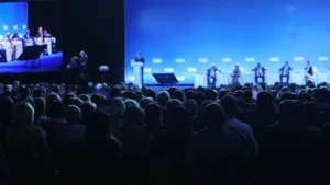 Large audience seated in a conference hall watching a panel discussion on a blue lit stage with speakers and a moderator