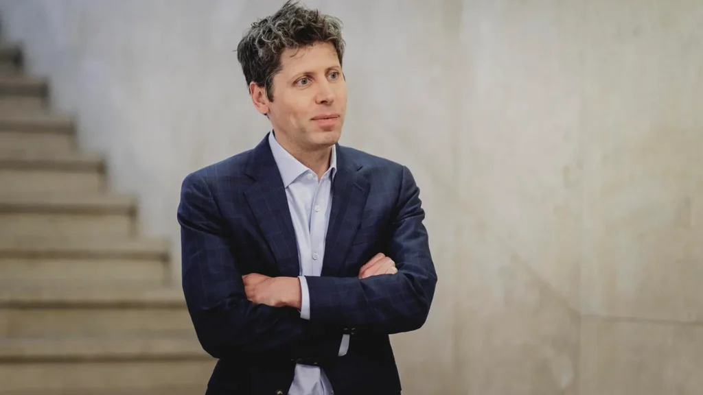 Sam Altman at a Technology and Policy Event | Brimco Sam altman wearing a dark blazer and light blue shirt stands with arms crossed in front of a neutral background