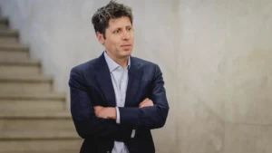 Sam altman wearing a dark blazer and light blue shirt stands with arms crossed in front of a neutral background