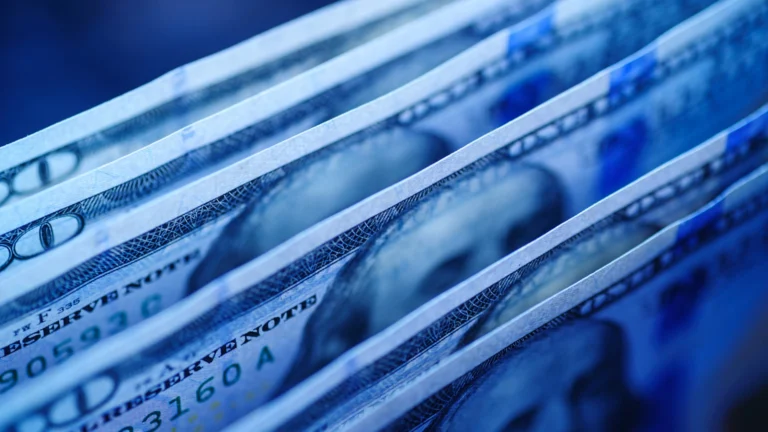 Close-up view of multiple U.S. dollar banknotes fanned out, highlighting details of $100 bills in cool blue lighting.