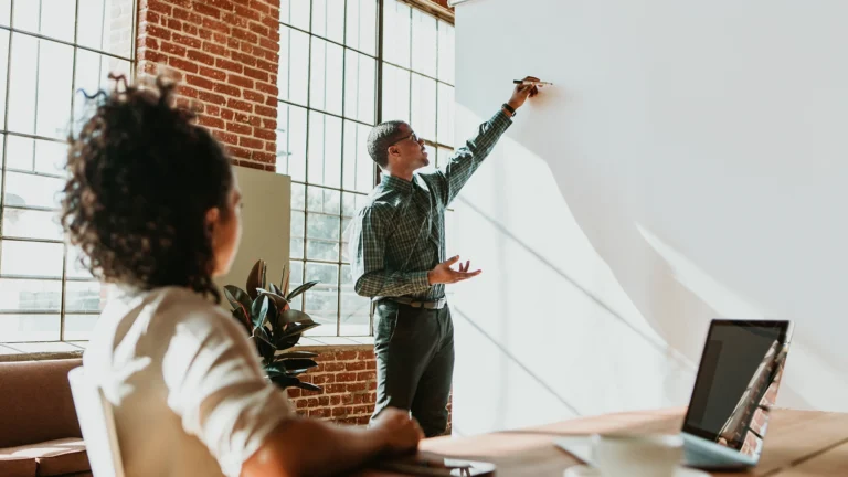 A man presenting ideas on a whiteboard during a meeting while a colleague listens at a table with a laptop in a modern office.
