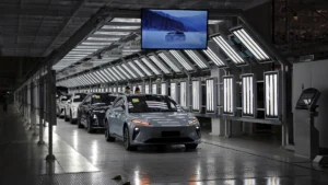 Electric vehicles move through a brightly lit automotive assembly line in a modern chinese factory with quality inspection stations and overhead displays
