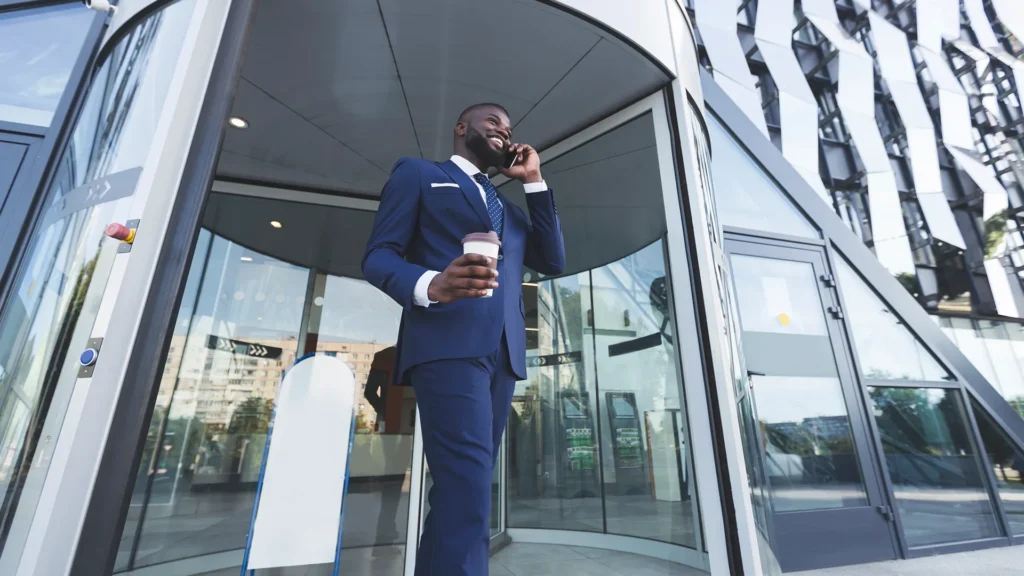 Successful businessman in blue suit walking out of modern office building while talking on phone and holding coffee.