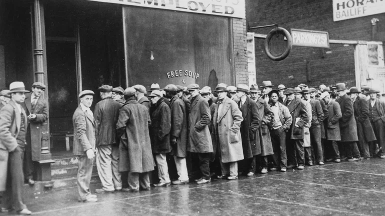A long line of unemployed men in coats and hats stands outside a building with a "Free Soup" sign during the Great Depression.