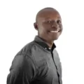 Smiling man in a gray button-down shirt, facing slightly to the side, against a white background.